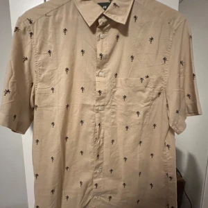 Beige skjorta med palmmönster från H&M - Snygg beige skjorta från H&M med korta ärmar och svart palmmönster över hela plagget. Skjortan har klassisk krage, bröstficka och knappar framtill. Perfekt för chill dagar och sommarhäng. Materialet är mjuk bomull som känns skönt mot huden.