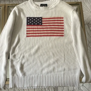 Vit stickad tröja med USA-flagga - Vit stickad tröja från Crocker i storlek S med en stor USA-flagga broderad på bröstet. Tröjan har rund hals, ribbade muddar och långa ärmar. Perfekt för dig som gillar klassisk stil med en cool twist.