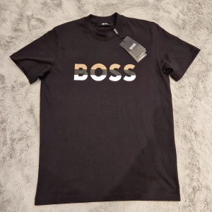 Svart BOSS t-shirt med tryck - Svart t-shirt från BOSS med stort, stilrent BOSS-tryck i beige, grått och vitt på bröstet. Klassisk rund hals och korta ärmar. Skön bomullskvalitet som passar perfekt till jeans eller shorts. Snygg och enkel design med modern vibe. Passar M och S