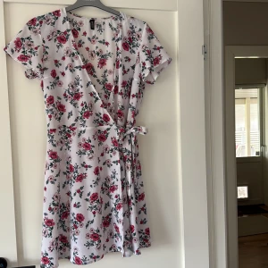 Blommig omlottklänning från H&M - Superfin kort omlottklänning från H&M i vitt med rosa och röda blommor. Klänningen har korta ärmar, knyt i midjan och v-ringning. 