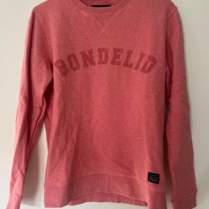 Rosa sweatshirt från Bondelid - Säljer en rosa sweatshirt från Bondelid med tryckt logga över bröstet. Tröjan har rund halsringning, ribbade muddar och är tillverkad i mjuk bomullsmix. Perfekt för en chill och avslappnad stil.