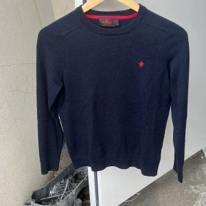 Mörkblå merinoullströja från Morris - Snygg mörkblå stickad tröja från Morris i 100% merinoull. Klassisk modell med rund halsringning och diskret röd broderad logga på bröstet. Perfekt för dig som gillar stilrena och tidlösa plagg med en modern touch. Jätte fint skick. Säljer kläder då de har blivit för litet.