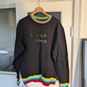 Golf svart tröja med regnbågsdetaljer - Svart långärmad tröja från Golf med broderad text 'CHERRY BOMB' i färgglada bokstäver på bröstet. Krage, ärmslut och nederkant har ränder i rött, gult, grönt och blått. På ena ärmen står det 'FIND YOUR WINGS' i gult. 100% bomull.