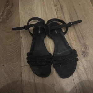 Svarta sandaler med flätade band - Svarta sandaler från Paceland med två flätade band över foten och smal rem runt ankeln. Snygg och enkel design med platt sula, perfekt för varma dagar. Passar till både jeans och klänning.