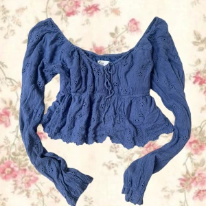 Blå broderad blus med snörning - Supersöt blå blus med broderade hålmönster och volangkant. Den har långa ärmar, v-ringning och snörning framtill. Perfekt för dig som gillar romantiska detaljer och en feminin stil.