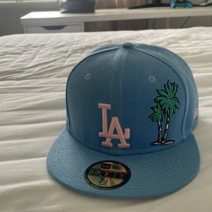Blå LA Dodgers keps från New Era - Säljer en blå 59FIFTY keps från New Era med LA Dodgers-logga i ljusrosa framtill och broderade palmträd. Skärmen är ljusrosa undertill och kepsen har broderad badge på sidan. Perfekt för dig som vill sticka ut med en färgglad och sportig stil.