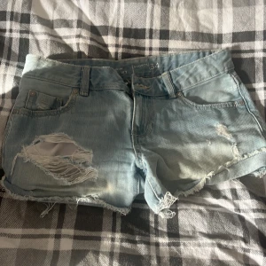 Ljusblå slitna jeansshorts - Säljer ett par ljusblå jeansshorts med råa kanter och slitningar framtill. Shortsen har klassisk femficksdesign, bälteshällor och en avslappnad vibe. Perfekta för varma dagar när du vill ha en cool och avslappnad look.