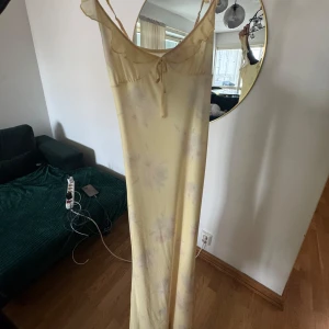 Ljusgul långklänning med volang - Superfin ljusgul långklänning med tunna axelband, volangdetaljer vid bysten och knyt framtill. Klänningen har ett subtilt blommigt mönster och är i ett lätt, flowy material som känns sommarfräscht och romantiskt.