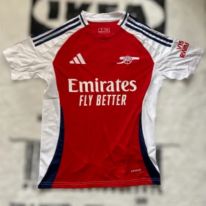 Arsenal hemmatröja Adidas röd/vit S  - Arsenal hemmatröja från Adidas i slim fit, tillverkad i lätt och ventilerande Aeroready-material. Klassisk röd front med vita ärmar och mörkblå detaljer på axlarna. Broderat Arsenal-emblem och Adidas-logga, samt sponsortryck på bröstet.