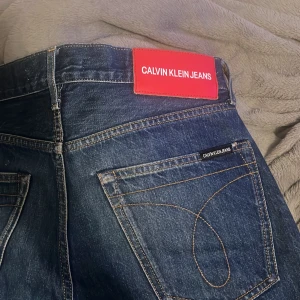 Calvin Klein Jeans mörkblå jeans - Mörkblå jeans från Calvin Klein Jeans med klassisk femficksdesign och snygga kontrastsömmar. Byxorna har en tydlig röd logotyppatch bak i midjan och subtil logga på bakfickan. Jeansen är raka i modellen och tillverkade i slitstarkt denim.