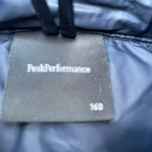 Blå dunjacka Peak Performance 160 - Snygg mörkblå dunjacka från Peak Performance med huva och dragkedja framtill. Jackan har två fickor med dragkedja och ribbade muddar vid ärmsluten. Perfekt för kalla dagar och riktigt skön att ha på sig.