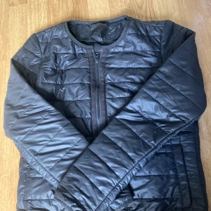 Svart quiltad pufferjacka från Vila - Svart pufferjacka från Vila i storlek S. Jackan är quiltad med horisontella sömmar, har rund halsringning och dragkedja framtill. Tillverkad i syntetmaterial och har långa ärmar samt praktiska fickor på sidorna. Perfekt för kyliga dagar nu till hösten.