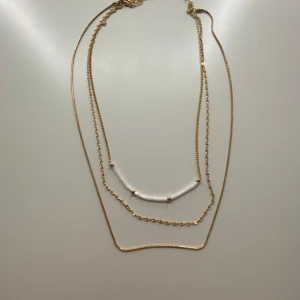 Guldigt lager-på-lager halsband - Snyggt lager-på-lager halsband med tre tunna kedjor i guldton. En av kedjorna har vita pärlliknande detaljer och små guldfärgade kulor. Perfekt för att ge din outfit lite extra edge och stil. Justerbart spänne för att passa olika längder.💕