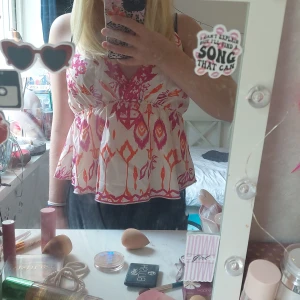 Färgglad blus med v-ringning - Säljer en söt blus med v-ringning och breda axelband. Blusen har ett färgstarkt mönster i rosa och orange på vit botten, med en flowy och luftig passform. Perfekt för dig som gillar boho vibes och vill sticka ut med coola färger och mönster.