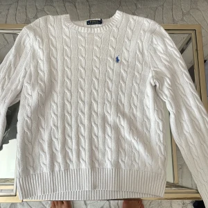 Vit kabelstickad tröja Polo Ralph Lauren - Säljer en vit kabelstickad tröja från Polo Ralph Lauren med klassisk blå logga på bröstet. Tröjan har rund hals, ribbade muddar och är långärmad. Perfekt för dig som gillar stilrena och tidlösa plagg med snygg struktur.