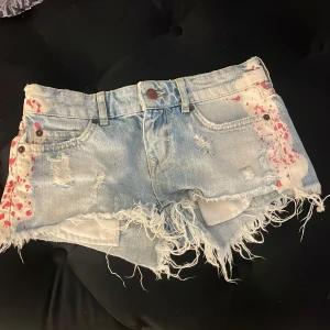 Ljusblå  jeansshorts Gina Tricot - Snygga ljusblå jeansshorts från Gina Tricot med råa, fransiga kanter och slitna detaljer. Shortsen har röda färgstänk på sidorna och klassisk femficksdesign. Perfekta för dig som gillar en edgy och unik stil.Dom är även low waist och finns inte att köpa i butik längre 
