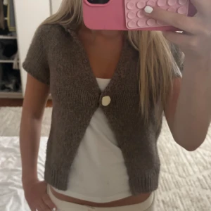 Brun stickad kofta med knapp - Kort brun stickad kofta med en stor vit knapp framtill. Koftan har korta ärmar och en djup v-ringning, perfekt att slänga över ett linne. Enkel och stilren design som passar till många outfits.