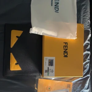 Fendi svart kuvertväska med gula detaljer - Säljer en svart kuvertväska från Fendi med ikoniska gula ögon-detaljer på framsidan. Väskan är tillverkad i skinn och har en stilren, rektangulär form. Kommer med originalbox, dustbag och äkthetsintyg. Perfekt för dig som vill sticka ut med en lyxig accessoar. Kan tänka mig att gå ner lite i pris vid en snabb och smidig affär😁