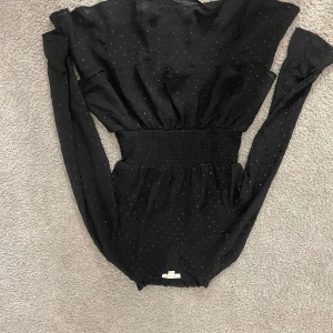 Svart prickig kort klänning med volang - Snygg svart kort klänning med små prickar och volangdetaljer vid axlarna. Klänningen har långärmade ärmar och en bred, smockad midja som ger en cool siluett. Perfekt för dig som gillar stilrena och trendiga plagg med lite extra detaljer.