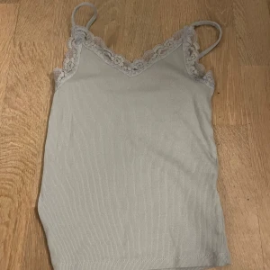 Ljusgrå ribbad linnetopp Primark XS - Supersöt ljusblå ribbad linnetopp från Primark med tunna axelband och spetsdetaljer längs v-ringningen. Perfekt att styla med jeans eller kjol för en chill look.