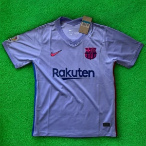 Lila FC Barcelona Nike fotbollströja - Snygg lila FC Barcelona fotbollströja från Nike med klubbmärke och Rakuten-tryck på bröstet. Kortärmad modell i lätt och ventilerande material, perfekt för match eller träning. La Liga-märke på ärmen och UNICEF-tryck på ryggen. V-ringad krage och diskreta blå/röda detaljer i sidorna.