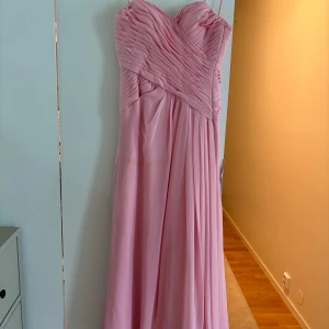 Lång rosa klänning med omlottliv - Säljer en elegant lång klänning i ljusrosa med omlottliv och plisserade detaljer upptill. Klänningen är axelbandslös och har ett svepande fall som ger en romantisk vibe. Perfekt för bal eller student! Materialet känns mjukt och har ett fint skimmer.