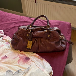  handväska från Marc Jacobs - Snygg trendig mörklila handväska från Marc Jacobs med guldfärgade metalldetaljer och en cool metallbricka framtill. Väskan är tidigare köpt på Plick och i bra begagnat skick. Men på baksidan har den några skrap märken där av ett lite lägre pris. Det långa bandet kommer även med. Hoppas den hittar ett nytt hem🤩🤩