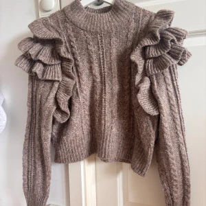 Brun stickad tröja med volang Gina Tricot - Säljer en brun  stickad tröja från Gina Tricot i storlek M. Tröjan har höga ribbade muddar, kabelstickat mönster och volanger på axlarna som ger en cool och unik look. Storlek m men kan upplevas lite mindre!