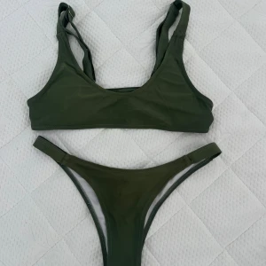 Olivgrön bikini med coola banddetaljer - Snygg olivgrön bikini med sportig topp och smala axelband som går att justera. Baksidan har unika banddetaljer som ger en edgy look. Underdelen är högt skuren och har smala sidor. Perfekt för dig som vill sticka ut på stranden i sommar!