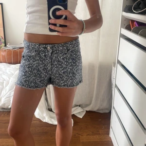 Blommiga jeansshorts från H&M - Snygga blå jeansshorts från H&M med vit blommigt mönster. Modellen är lågmidjad med klassisk femficksdesign, rå kant nedtill och dragkedja med knapp. Perfekta för varma sommardagar och har en fräsch, ungdomlig vibe.