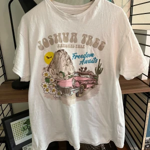 Amerikansk  vintage-t-shirt med tryck - Amerikans vintage-T-shirt från National Park Foundation i 100% bomull. Framsidan har ett färgglatt tryck med texten 'Joshua Tree National Park', blommor, berg, kaktus och en rosa retrobil. Baksidan har samma text i beige. Klassisk passform och rund hals.