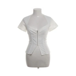 Vit korsett-topp med dragkedja - Snygg vit korsett-topp med korta ärmar och markerad midja. Toppen har en v-ringad hals och en dragkedja framtill som ger en edgy look. Croppad passform som visar lite mage. Perfekt att styla med jeans eller kjol för en trendig outfit.