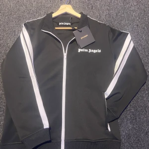 Svart Palm Angels track jacket S - Svart track jacket från Palm Angels med vita ränder längs ärmarna och logga på bröstet. Jackan har hel dragkedja framtill, ribbade muddar och sportig krage. Tillverkad i 100% polyester för en skön och lätt känsla. Perfekt för en streetwear-look. Helt ny med prislapp, inget kvitto😃