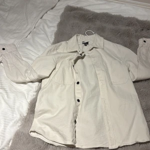 Beige overshirt  XS - Snygg beige overshirt från H&M i storlek XS. Skjortan har två bröstfickor, svarta knappar och klassisk krage. Materialet är mjukt och känns som bomull, perfekt att ha som lager. Långärmad och stilren design som funkar till många outfits.