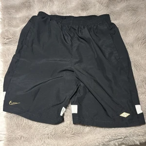 Svarta Nike Dri-Fit shorts  - Svarta Nike Dri-Fit shorts i storlek XL med vita meshdetaljer på benen och guldfärgad Nike-logga. Elastisk midja för skön passform, tillverkade i lätt syntetmaterial som andas. Perfekta för sport och träning, stilrena och funktionella.