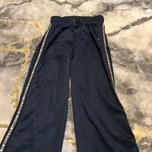 Svarta wide mjukisbyxor Tommy Hilfiger - Svarta mjukisbyxor från Tommy Hilfiger i wide modell med hög midja och snörning i midjan. Snygga logoband längs sidorna och liten broderad flagga på ena benet. Sköna och stilrena, perfekta för chill eller streetwear. Materialet är mjukt och känns riktigt bekvämt.