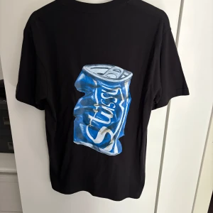 Svart Stüssy t-shirt med blå burk - Svart t-shirt från Stüssy i bomull med coolt tryck av en blå krossad läskburk på ryggen och Stüssy-logga på bröstet. Klassisk rund hals och korta ärmar. 