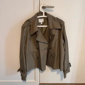 Grön trenchcoat - Snygg grön trenchcoat från H&M med klassisk krage, dubbelknäppning och bälte vid ärmsluten. Knappt använd och perfekt inför hösten