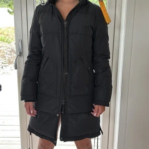 Svart Parajumpers dunparkas med päls - Säljer en svart lång dunparkas från Parajumpers med päls på huvan och ikonisk gul rem med metallspänne vid axeln. Jackan har två stora fickor fram, dragkedja och broderad patch på ärmen. Perfekt för kalla dagar och riktigt snygg siluett. Dragkejan är inte original utan utbytt mot en svart som inte fastnar.