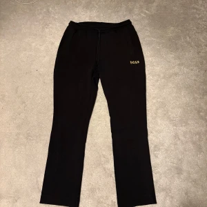 Svarta mjukisbyxor från BOSS - Svarta mjukisbyxor från BOSS med gul logotyp broderad på vänster ficka. Byxorna har elastisk midja med snörning och raka ben. Perfekta för chill eller streetwear, med mjukt bomullsmaterial och praktiska sidofickor.