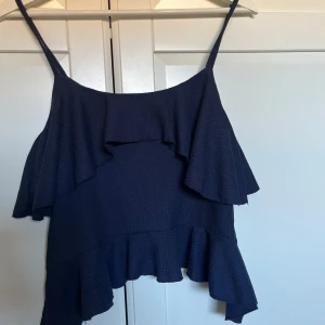 Mörkblå volangtop från Nelly XS - Supersöt mörkblå linnetopp från Nelly i storlek XS. Toppen har tunna axelband och flera lager med volanger som ger en flowig och chill vibe. Materialet är lätt och har en diskret struktur som gör den extra snygg. Perfekt att styla med jeans eller kjol.