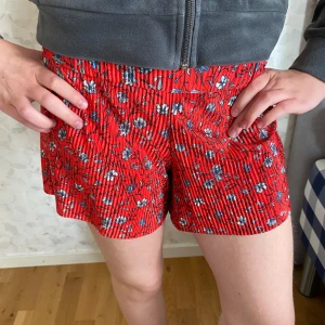 Röda blommiga shorts Gina Tricot XS - Snygga röda shorts från Gina Tricot i storlek XS med blommigt mönster i blått och vitt. De har en högmidjad passform och är gjorda i mjuk, plisserad polyester som ger en skön känsla. Perfekta för varma dagar när du vill sticka ut med färg och mönster.