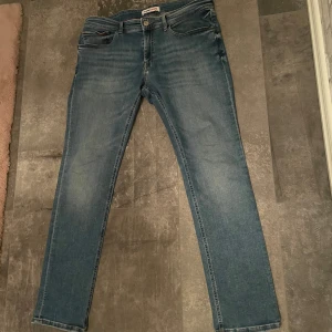 Tommy Jeans blå jeans, storlek 36/32 - Säljer ett par snygga blå jeans från Tommy Jeans i storlek 36/32. De har klassisk femficksdesign, raka ben. Jeansen har röda sömdetaljer och en patch med logga bak i midjan. Perfekta för dig som gillar stilrena och tidlösa jeans.