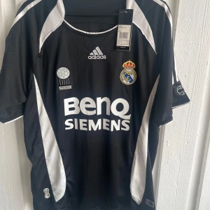 Real Madrid Robinho #10 svart matchtröja - Snygg svart Real Madrid fotbollströja från Adidas med vita detaljer och tryck. Tröjan har korta ärmar, klubbmärke på bröstet, Champions League-märke på ärmen och Robinho #10 på ryggen. Materialet är lätt och andas, perfekt för match eller träning.