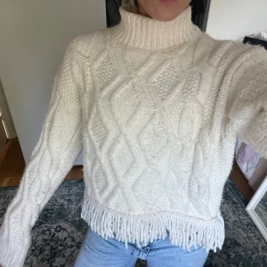 Vit polotröja med fransar - Säljer en chunky vit polotröja med grovt stickat kabelmönster och långa ärmar. Tröjan har hög krage och coola fransdetaljer längst ner, vilket ger en unik vibe. Perfekt att styla med jeans för en chill look.