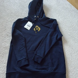 Marinblå oversized hoodie med broderi - Den ör helt ny, aldrig använd med lappar kvar, ordinarie pris 1099. Köpt på herravdelningen, men ser att den marknadsförs som unisex. Stor i storlek. marinblå oversized hoodie från SikSilk med snyggt broderat guldemblem på bröstet. Tröjan har huva med snörning, ribbade muddar och stor ficka framtill. Perfekt för chill dagar och streetstyle. Materialet är mjuk bomull.