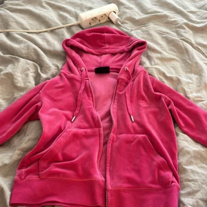 Juicy Couture rosa velour hoodie - Juicy Couture hoodie i stark rosa velour med broderad JC-logga på bröstet. Tröjan har huva med snörning, dragkedja framtill och två stora fickor. Mjukt material och klassisk juicy-stil, perfekt för chill eller streetwear. Metalländar på snöret med Juicy Couture-gravyr. Storleken på tröjan är S och byxorna XXS, säljer hela sättet för 800kr. Dock har själva ”J” på tröjan åkt av i torktumlaren och går inte att få på😔Vid frågar skriv till mig, så svarar jag så fort som möjligt! Mvh Elina💗