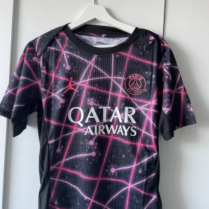 PSG x Jordan svart och rosa fotbollströja - Snygg PSG x Jordan fotbollströja i svart med rosa grafiskt mönster och vita Qatar Airways-tryck. Kortärmad med rund hals och tillverkad i lätt, ventilerande polyester. Paris Saint-Germain-logga och Jumpman-logga på bröstet. Perfekt för dig som vill sticka ut på planen.