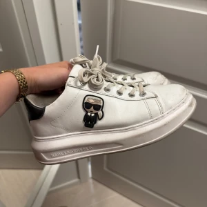 Karl Lagerfeld vita sneakers med ikon - Snygga vita sneakers från Karl Lagerfeld med coolt Karl-ikon på sidan och svarta detaljer vid hälen. Skorna har snörning, rund tå och platt sula. Tillverkade i skinn med metallfärgade öljetter och tjockare sula för extra stilpoäng.