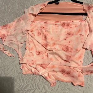 Blommig omlottopp från H&M - Säljer en superfin omlottopp från shein i ljusrosa mesh med blommigt mönster. Toppen har långa ärmar, v-ringning och knytband i midjan. Perfekt för dig som gillar romantiska vibes och vill ha något lätt och luftigt.